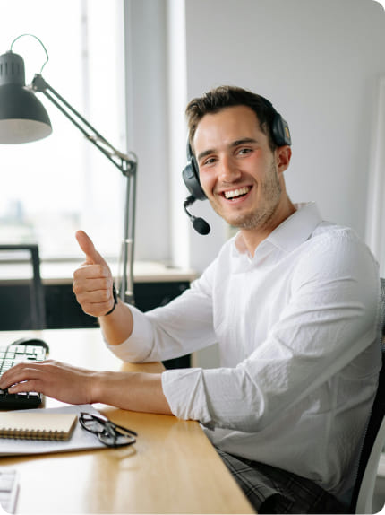 A man wearing a headset sits at a desk, smiling and giving a thumbs-up gesture while working on a computer in an office setting.