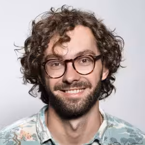 A man with curly brown hair, a beard, and round glasses is smiling at the camera. He is wearing a patterned shirt and is posed against a plain light background.